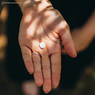 A close-up of an open hand holding a single round white pill outdoors, with sunlight and shadows on the skin.
