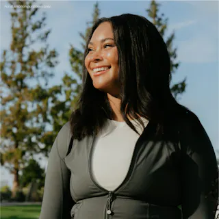 A woman wearing a gray jacket and white shirt stands outside, smiling, with trees and blue sky in the background.