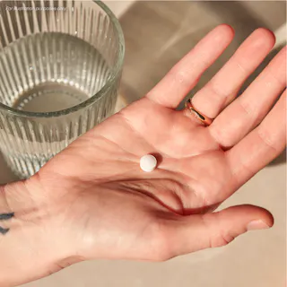 A hand holding a single white round pill with a glass of water in the background.