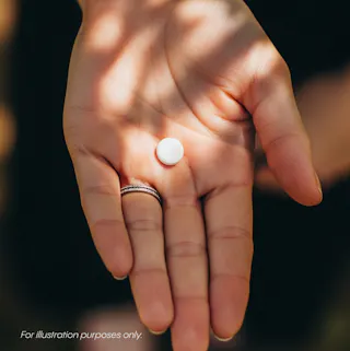 A hand holding a single round white pill, photographed in natural light. Text at the bottom reads, 