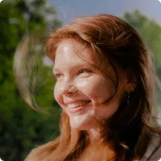 A young woman with red hair smiles outdoors, with sunlight on her face and greenery blurred in the background.